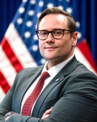 Image of John Marck in front of flag wearing a suit